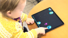 A young child at a table in a yellow shirt pointing a finger at a bubble on a tablet screen 