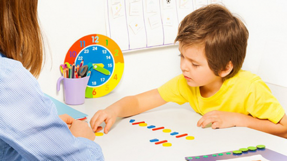A boy in a yellow shirt plays with colored blocks on a white table while a woman watches