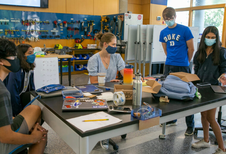 four students with masks stand around a work table in a tool-filled room