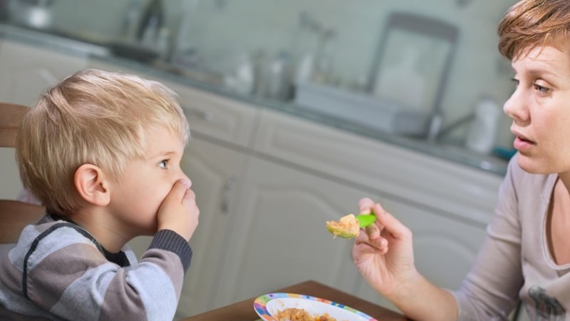 a child covers their mouth with a hand while an adult holds a spoon with food