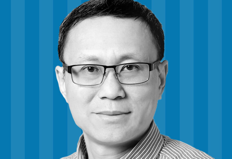 A black and white photo of a man with glasses and short hair in front of a blue striped background