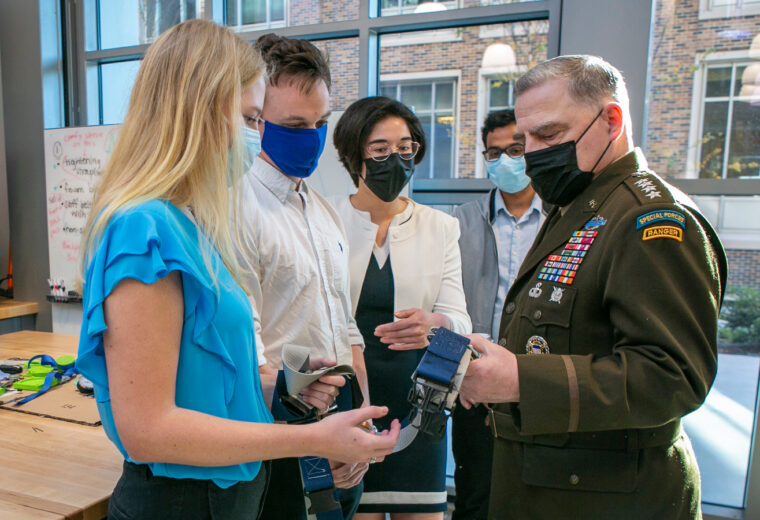 A man in a general jacket speaks with a team of students holding a belt-like object
