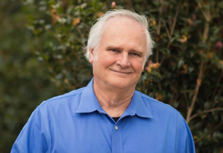 Man with white hair and blue shirt smiling in front of a tree