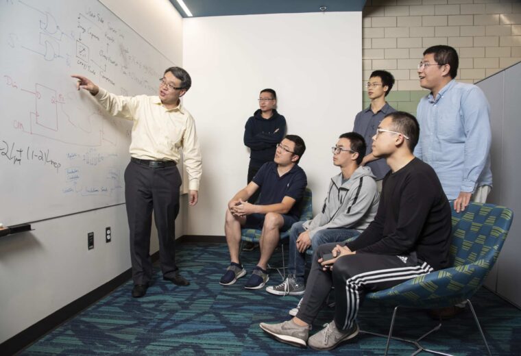 Yiran Chen points on a whiteboard as six graduate students observe.