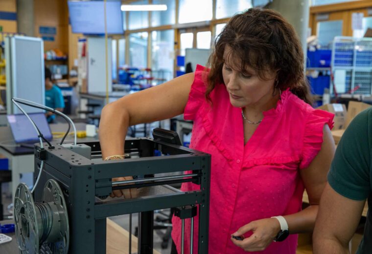 A woman wearing a pink blouse reaches her hand into a 3D printer.