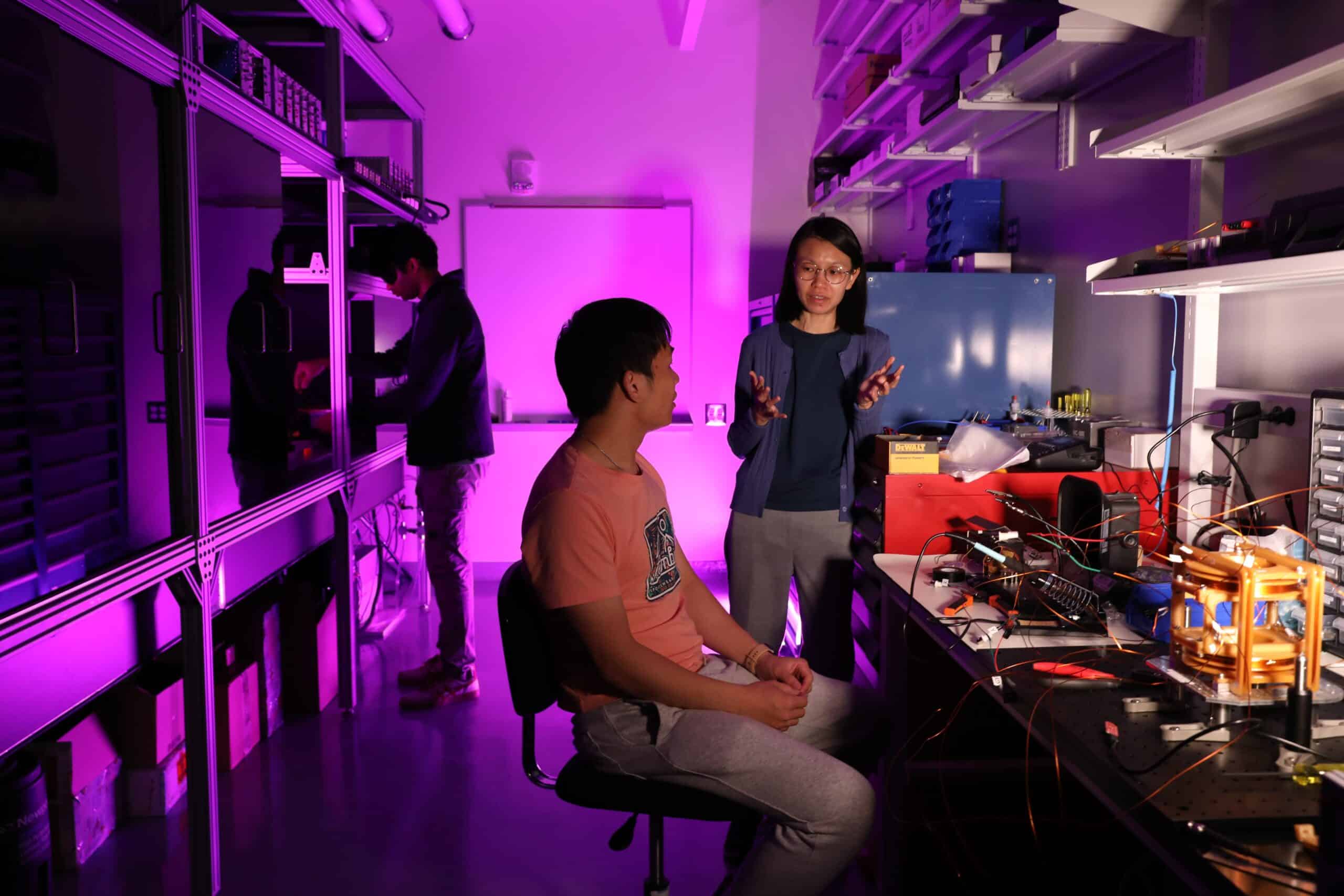 two graduate students and a faculty member work in a lab at the Duke Quantum Center, lit in purple
