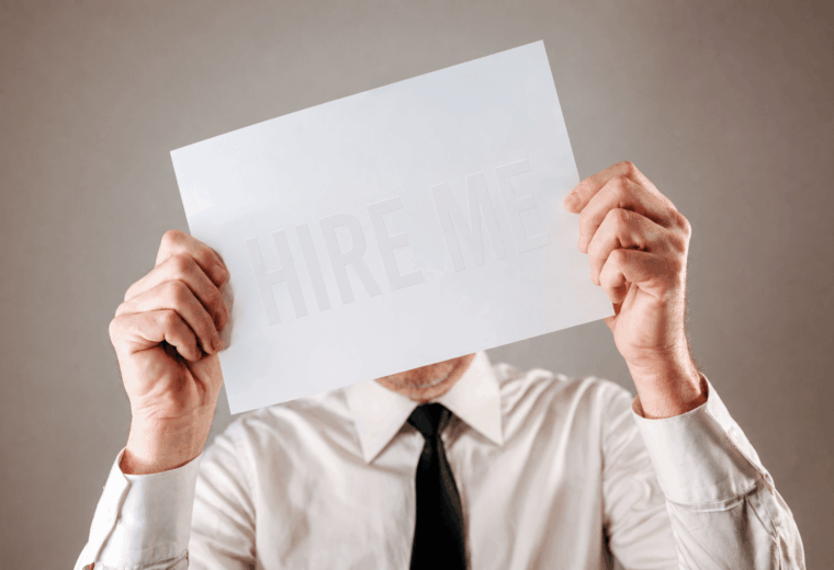 A man in a white shirt and black tie holding a white piece of paper in front of his face. The words "HIRE ME" are barely visible on the paper.